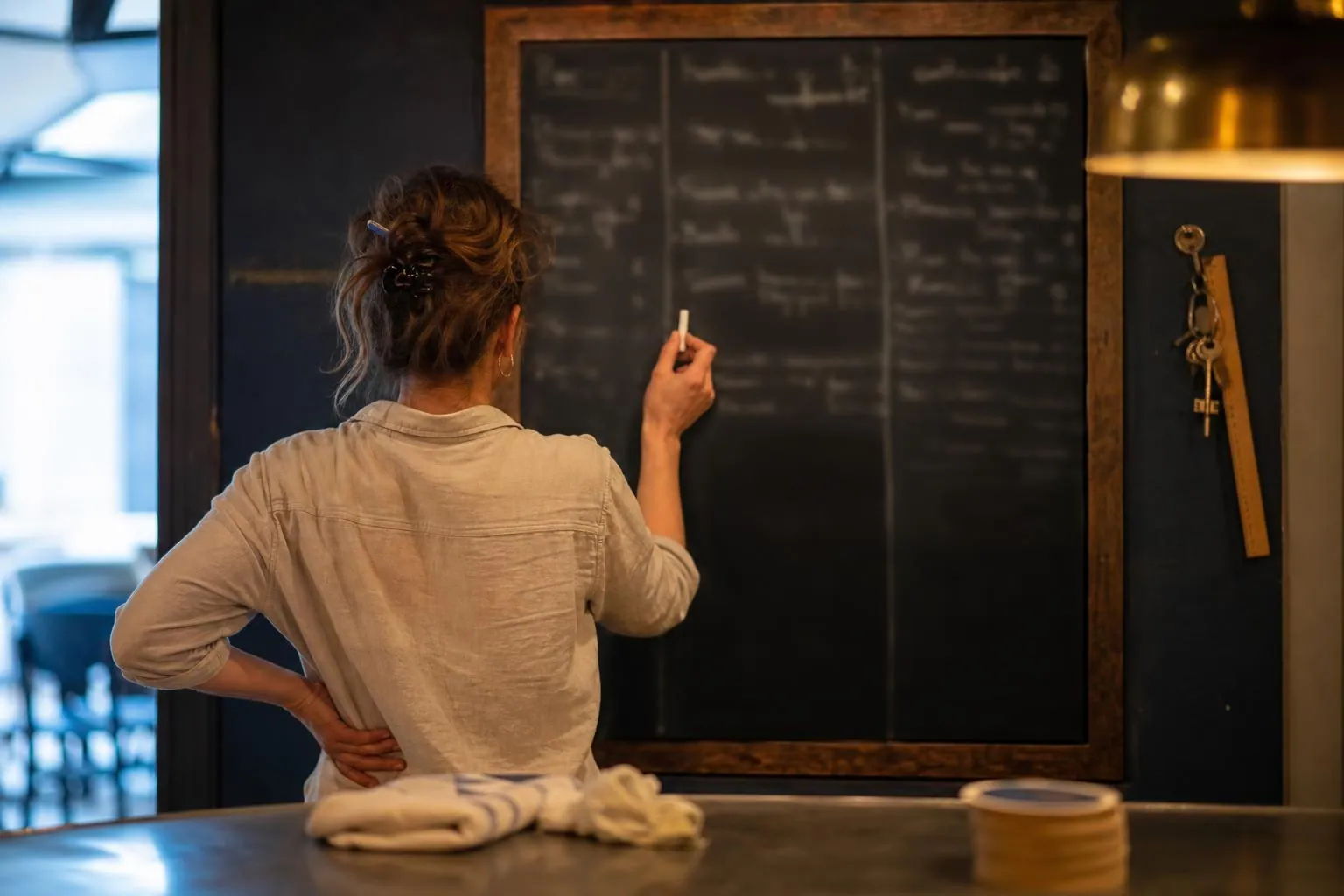 Main tenant une craie devant une ardoise murale de programmation derriere un comptoir de bar, lignes tracees et cadre en bois