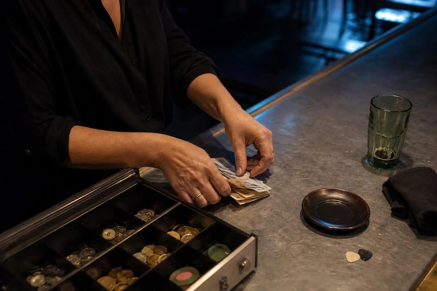 Mains d’une gérante comptant le fond de caisse en fin de soirée sur un comptoir en zinc, tiroir caisse ouvert et jetons à côté
