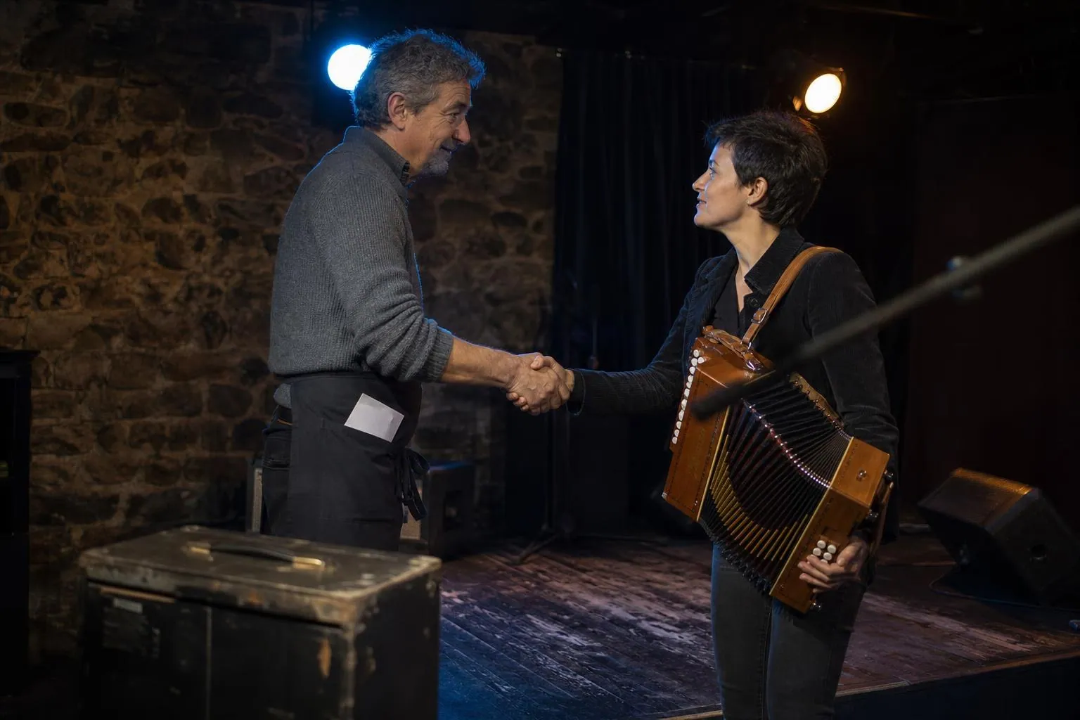 Poignée de main entre un exploitant et une accordéoniste au bord d’une petite scène, ampli et pied de micro autour d’eux