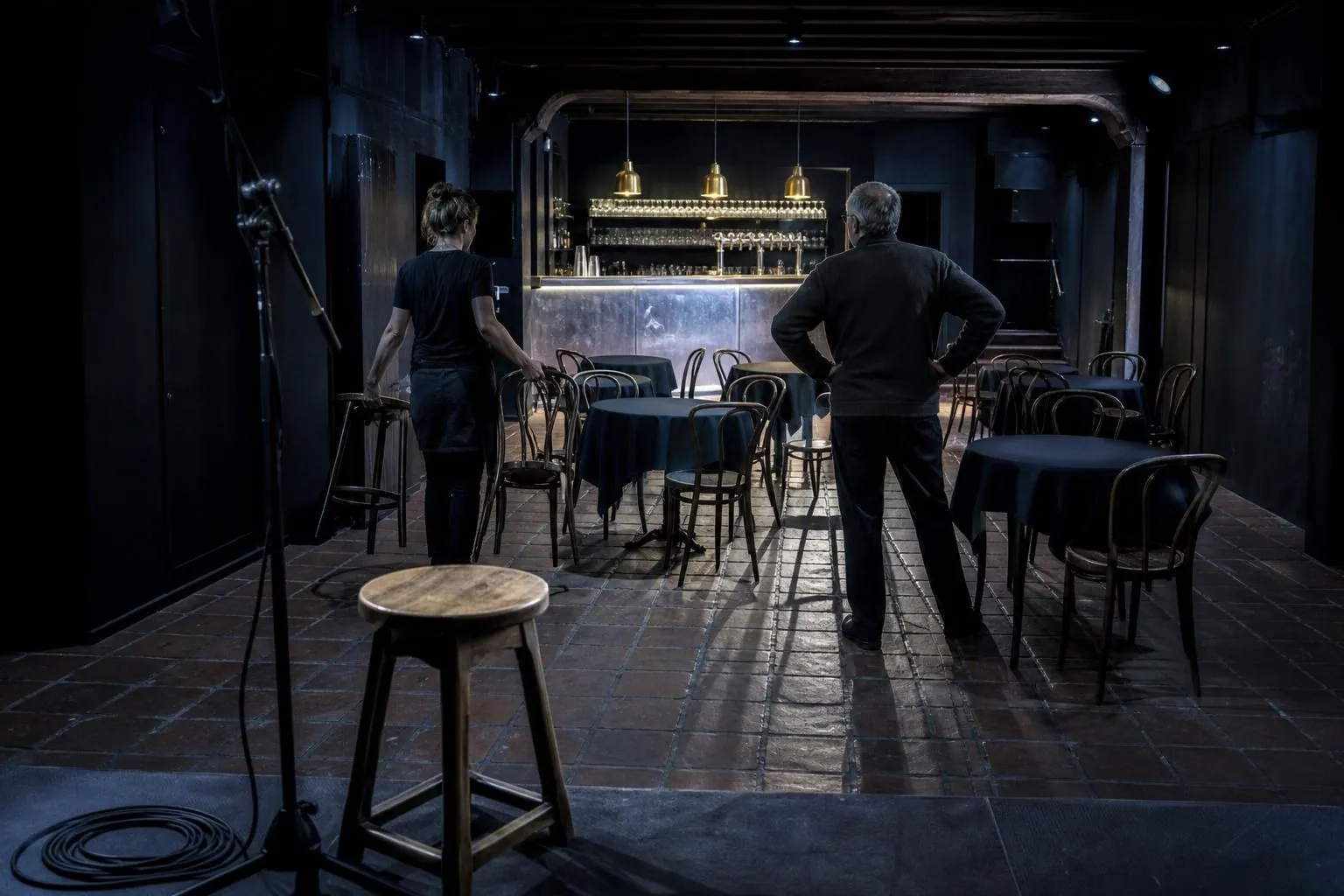 Salle de cafe concert vue depuis la scene avant l ouverture, tables dressees et deux personnes installant des chaises